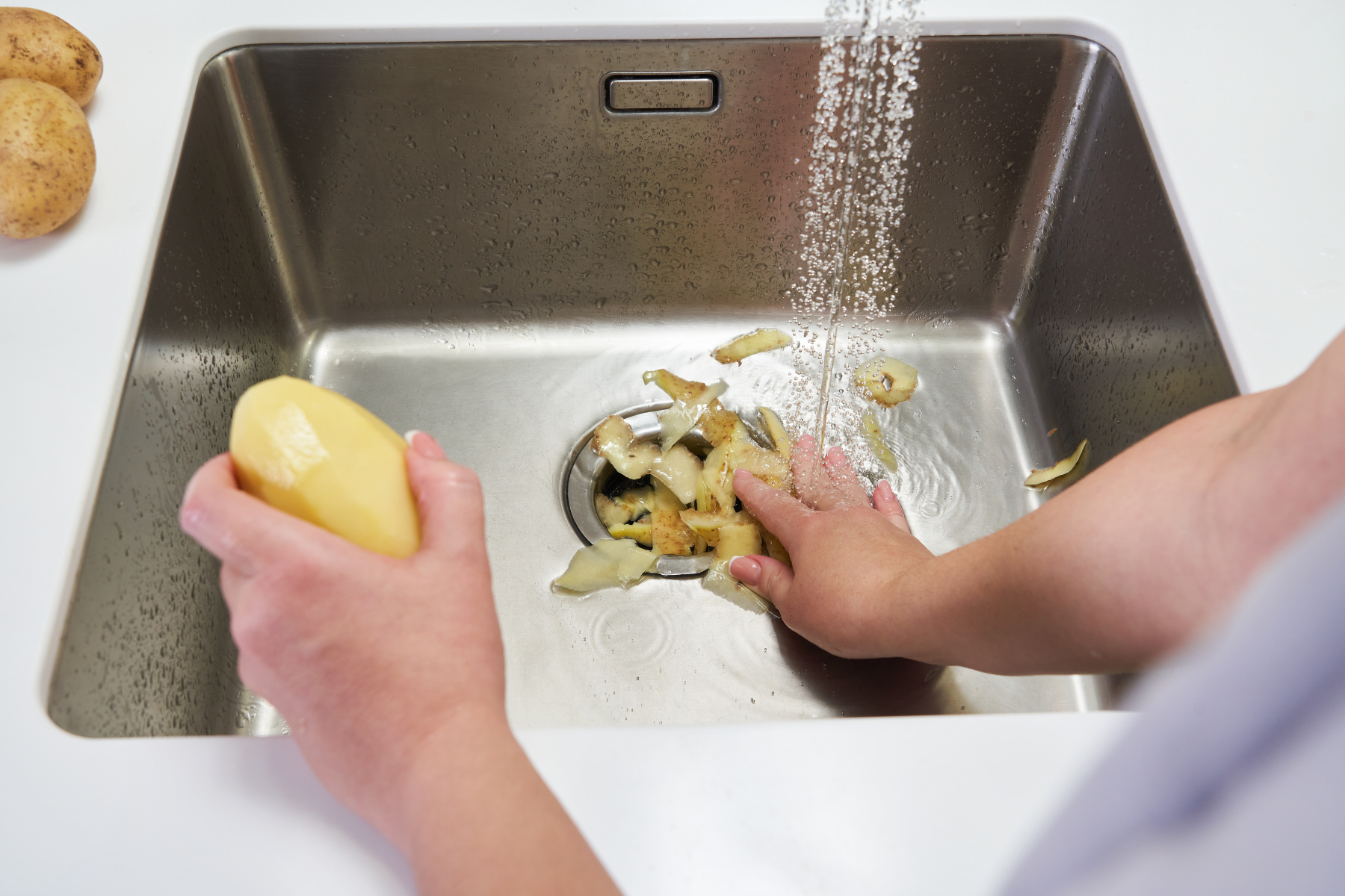 A person rinses potato peels down a kitchen sink drain with a garbage disposal while holding a peeled potato under running water.