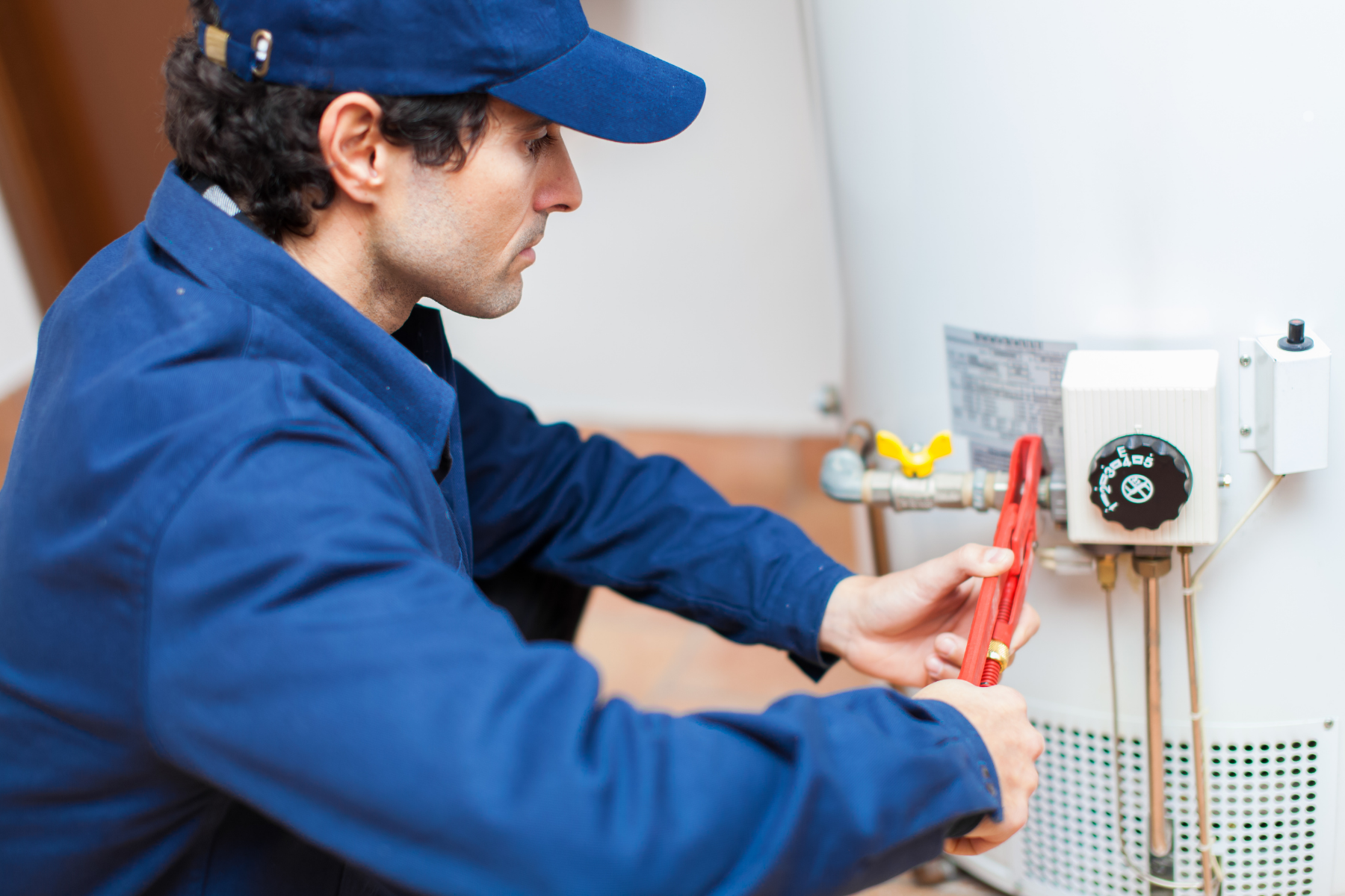 A technician in blue uniform and cap uses a red pipe wrench to adjust a fitting on an efficient gas water heater.