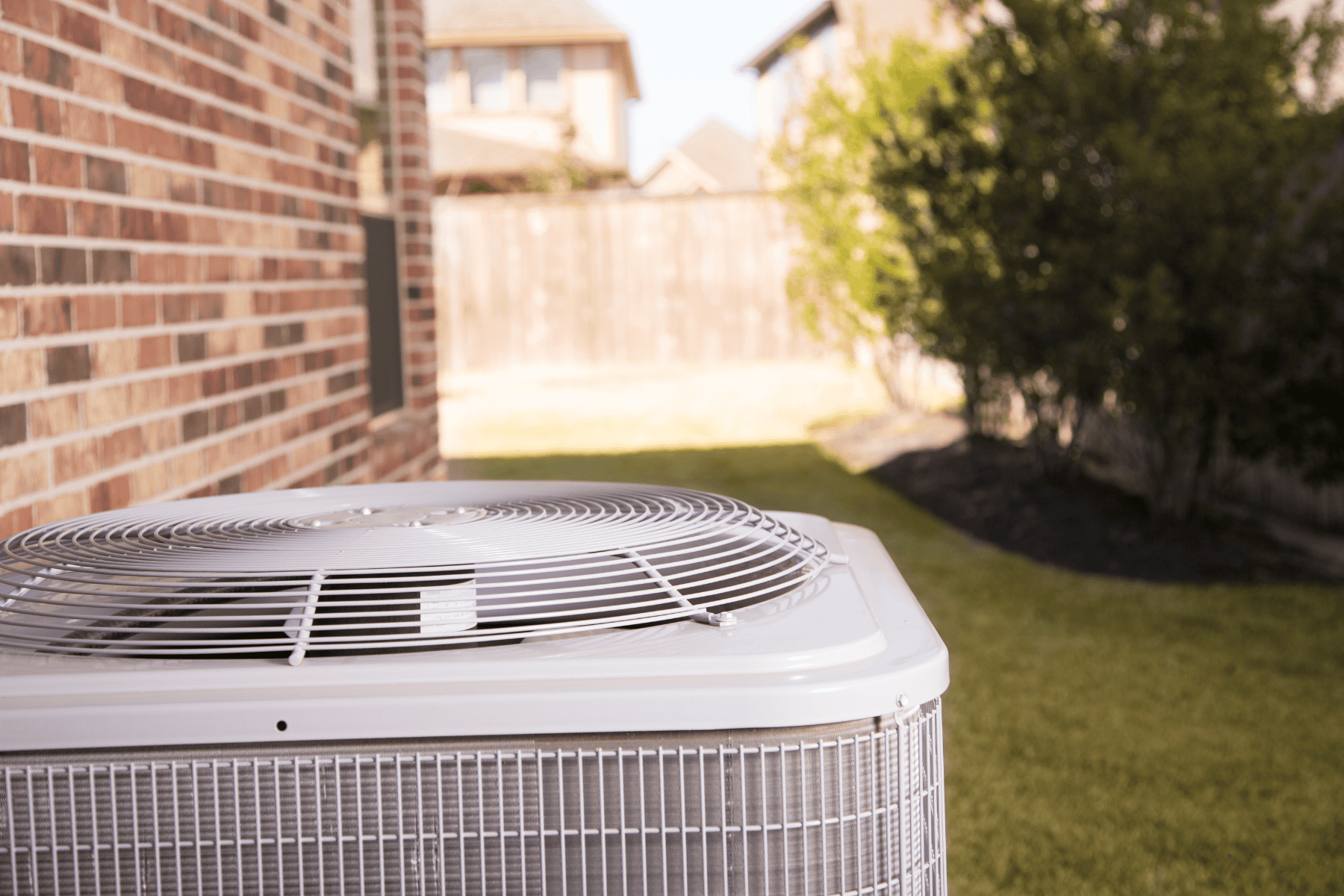 An outdoor central air conditioning unit sits beside a brick house on a grassy yard, with trees and a wooden fence in the background, showcasing an efficient forced air system to keep the home cool.