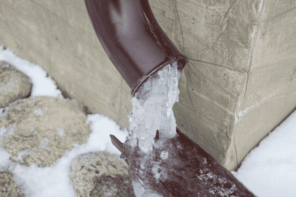 A brown downspout with ice blocking the opening, attached to a concrete wall, illustrates the importance of frozen pipes prevention. Snow and stones are on the ground around the area, serving as a reminder of how to prevent frozen pipes during harsh winter conditions.