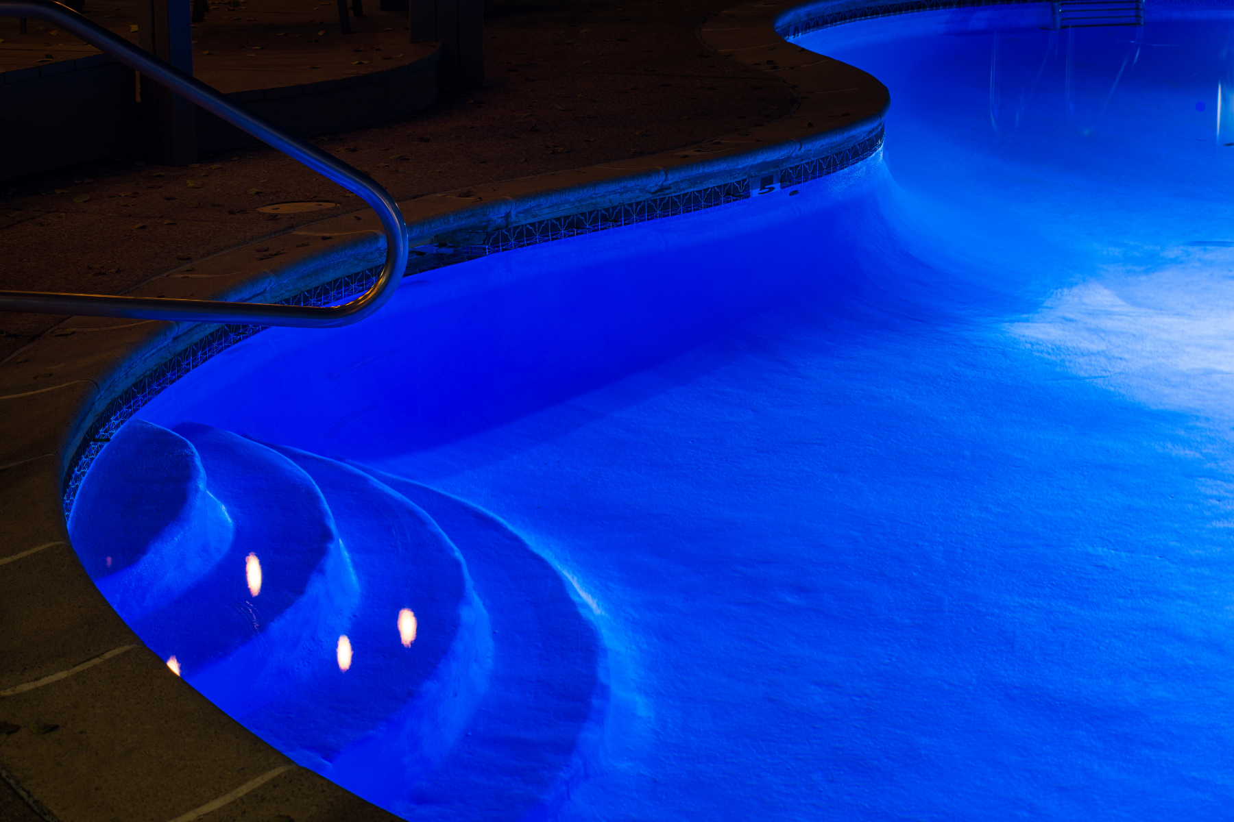 A well-lit pool at night, featuring bright blue underwater lights that illuminate curved steps and part of the deck—an inspiring option for stylish pool lighting.