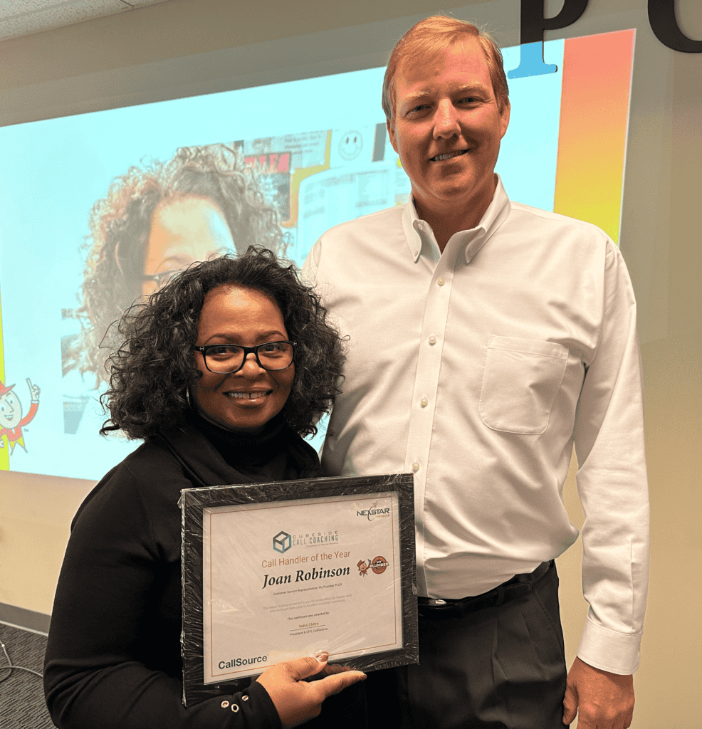 Two people stand indoors; the woman on the left holds a framed “Call Handler Award: Call Handler of the Year, Joan Robinson” certificate. A projected My Plumber Plus Customer Service Excellence presentation is visible in the background.