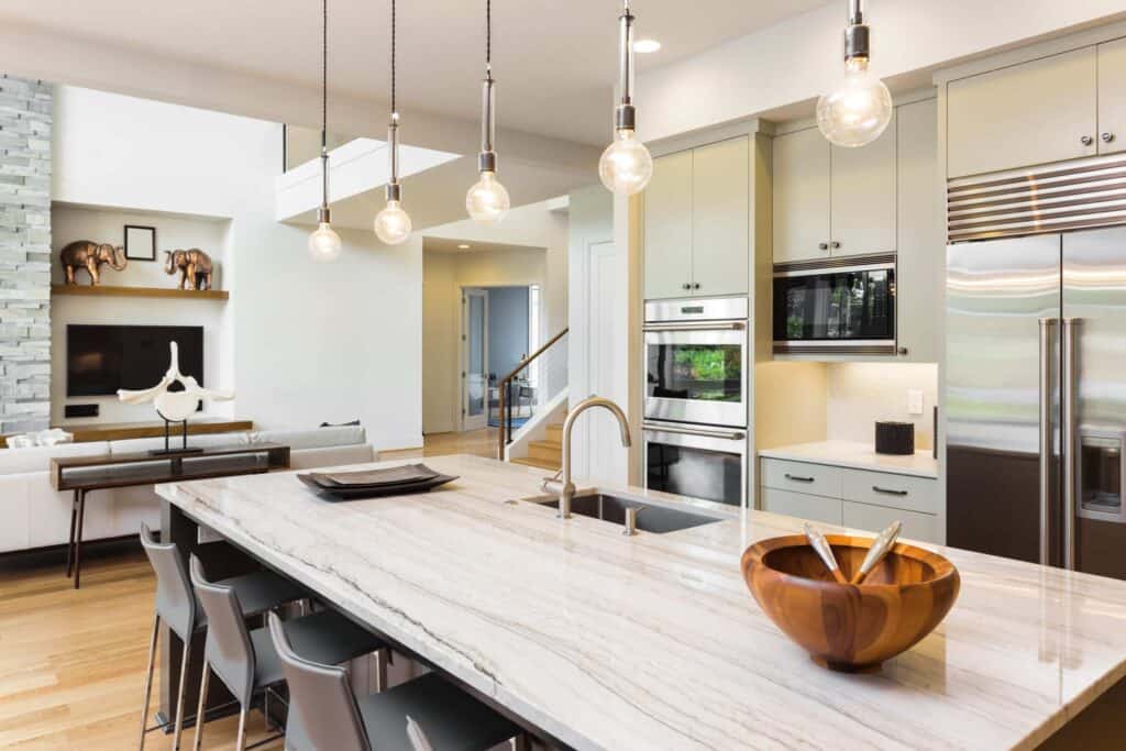 Modern kitchen with a marble island, pendant lights, stainless steel appliances, and stools—perfect for keeping it clean—plus a living area and staircase visible in the background.