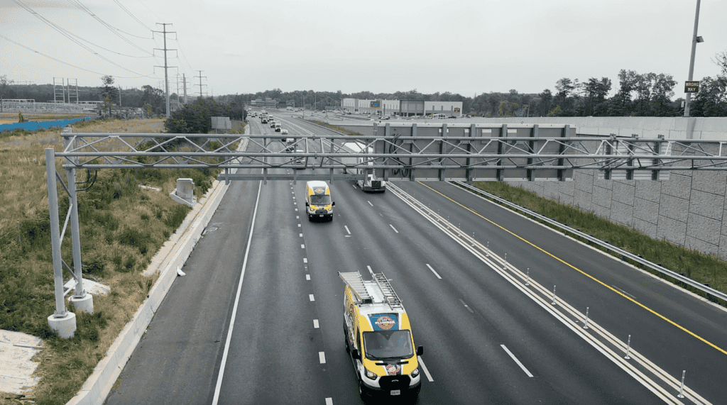 A wide highway with multiple lanes, light traffic, and several yellow My Plumber Plus service vans traveling in one direction under a metal overhead structure.