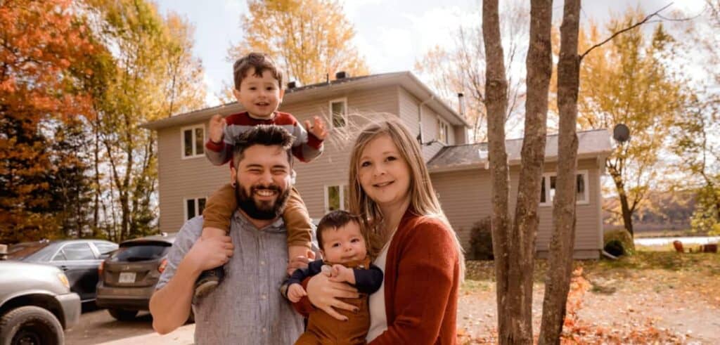A smiling family of four poses outside in front of a house with autumn trees in the background, capturing one of those pivotal moments that echo the warmth and unity seen throughout American history. The father holds a child on his shoulders; the mother holds a baby.
