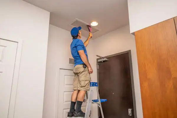 A person standing on a step ladder cleans an air vent on the ceiling inside a room, demonstrating professional air duct cleaning in Fairfax VA.