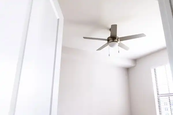 A modern ceiling fan with five blades is mounted on a white ceiling in a bright, minimal room with sunlight streaming through window blinds after expert ceiling fan installation in Fairfax VA.