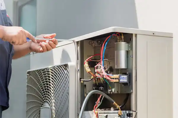 A person uses a screwdriver to repair or maintain an outdoor air conditioning unit with exposed wiring and components, similar to services provided for furnace repair in Fairfax VA.