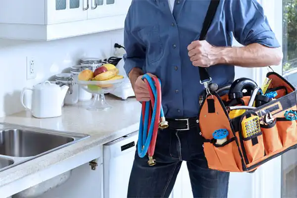 A person in a blue shirt stands in a kitchen in Fairfax VA, holding an orange tool bag and colored hoses, ready for kitchen plumbing repair near a counter with fruit and a sink.