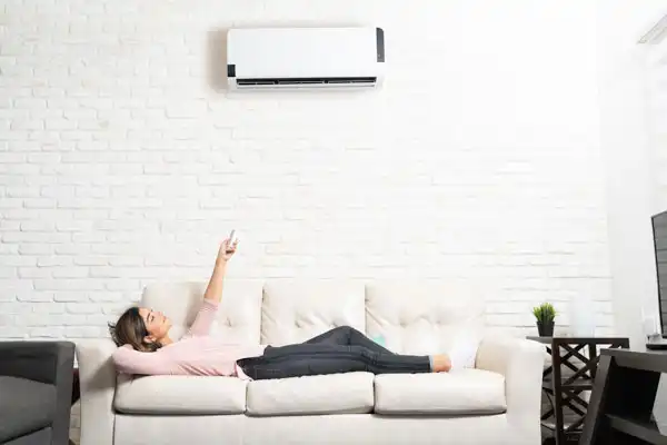 A person lying on a white sofa points a remote control at a wall-mounted air conditioner, enjoying the comfort from a recent mini-split installation in their bright Fairfax VA living room.