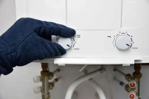A gloved hand adjusts the temperature dial on a white gas water heater in Fairfax VA, with visible pipes and control knobs labeled "Low" and "High," highlighting the importance of regular water heater maintenance.