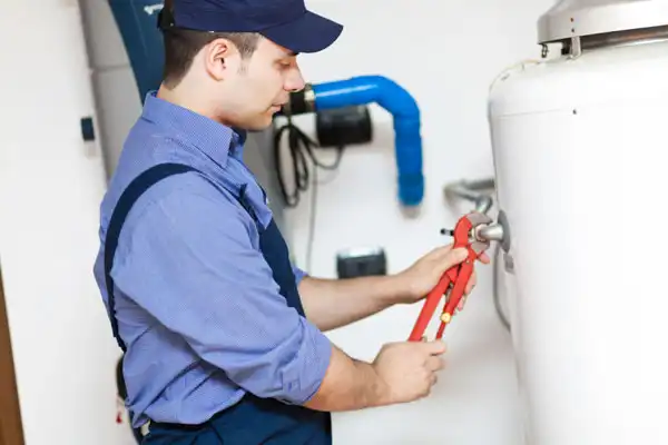 A technician in a blue uniform uses a wrench to adjust a valve on a white water heater in a utility room, performing water heater maintenance in Fairfax VA.
