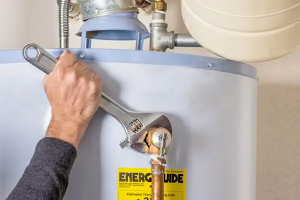 A person uses a large adjustable wrench to tighten a fitting on a water heater during a Water Heater Repair in Fairfax VA, with an Energy Guide label visible on the front.