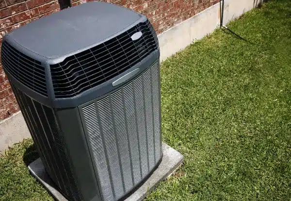 An outdoor air conditioning unit sits on a concrete pad next to a brick wall, surrounded by green grass—typical of expert Air Conditioning Installation in Fairfax, VA.