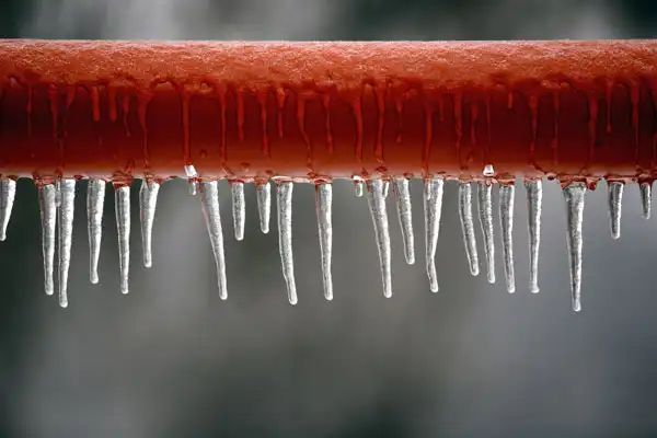 A row of icicles hangs from a frost-covered red metal pipe, hinting at the need for frozen pipe repair in Fairfax VA, set against a blurred gray background.