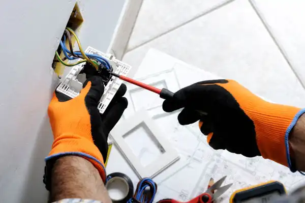 A person wearing orange and black gloves uses a screwdriver to perform Electrical Repair Fairfax VA, installing or repairing an electrical outlet on a wall.