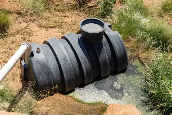 A black plastic septic tank, partially buried in the ground with a white pipe connected, sits surrounded by grass and muddy water—typical of septic tank repair sites in Fairfax, VA.