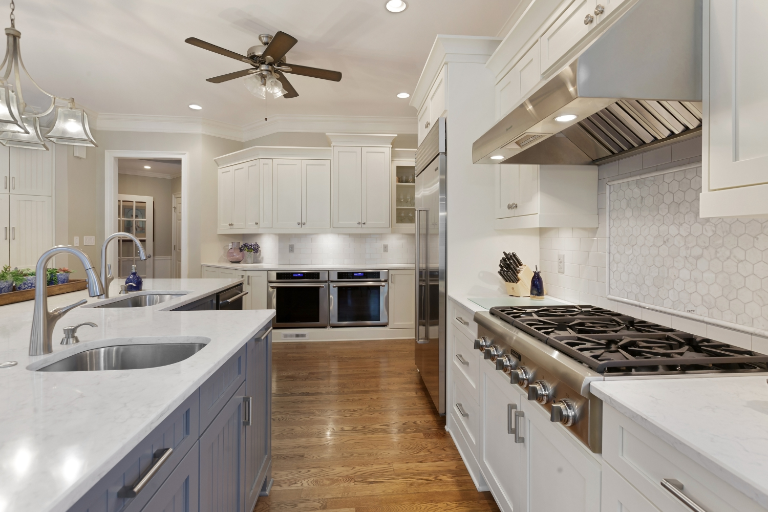 Modern kitchen with white cabinets, stainless steel appliances, double oven, gas stove, marble countertops, and wooden floors. Ceiling fan and pendant lights are visible—perfect for those who value safety and seek reliable home surge solutions.