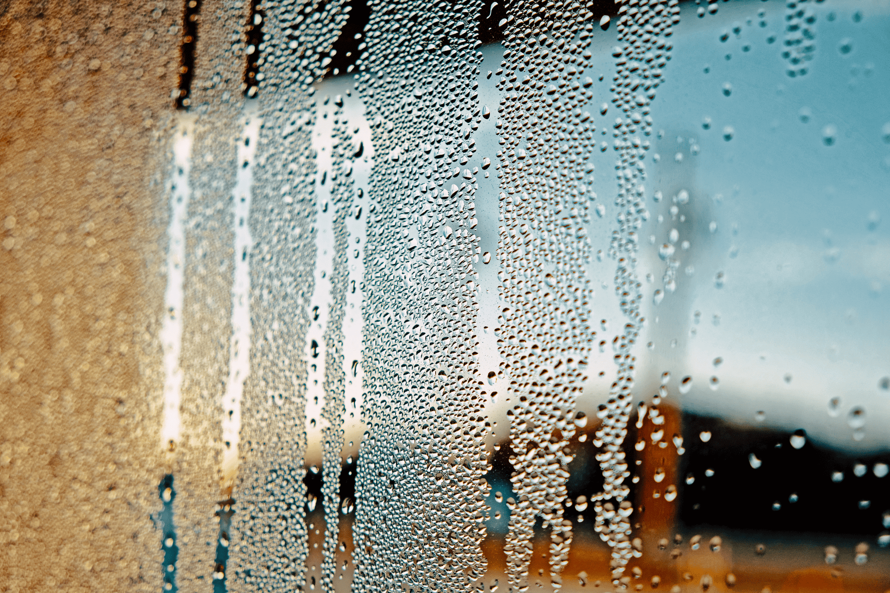 Condensation on a glass window with streaks and water droplets, set against an out-of-focus outdoor background in natural light—a common scene in any home, highlighting the need to choose a dehumidifier for improved comfort.