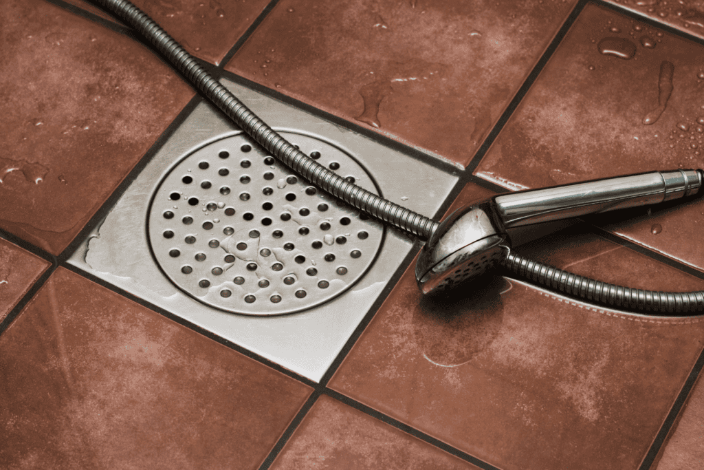 A handheld showerhead with a metal hose rests on a wet brown tiled floor next to a round metal drain cover, suggesting the lingering issue of a smelly shower drain.