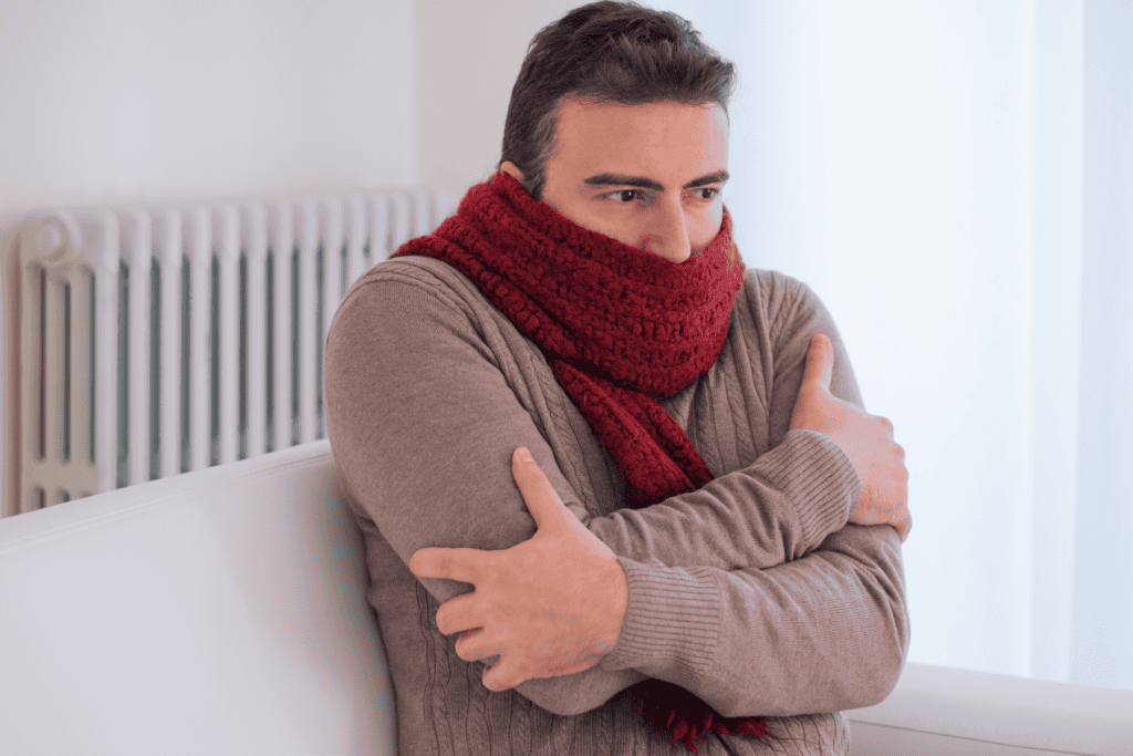 Man wearing a thick red scarf and beige sweater sits indoors with arms crossed, appearing cold. A white radiator is visible in the background—a familiar scene for many homeowners when the Emergency Heat or Aux Heat kicks in during chilly weather.