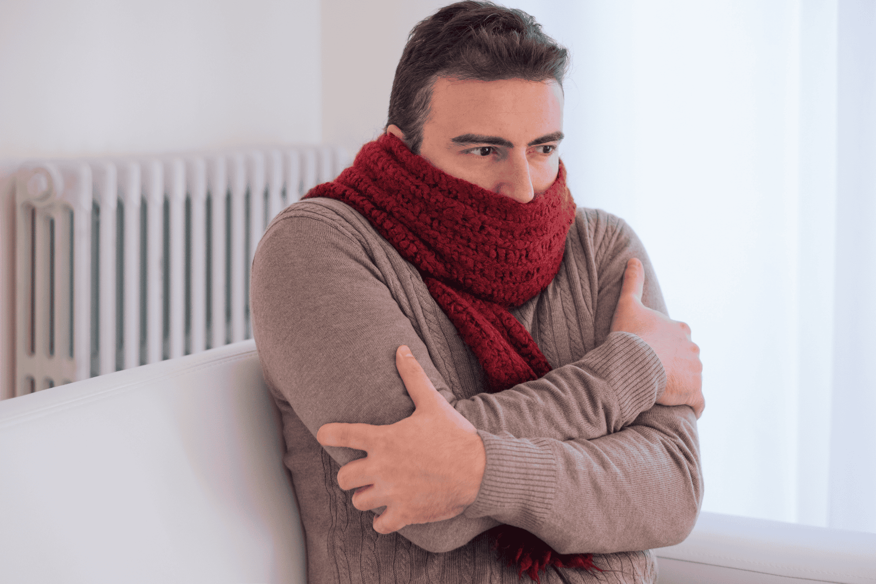 Man wearing a thick red scarf and beige sweater sits indoors with arms crossed, appearing cold. A white radiator is visible in the background—a familiar scene for many homeowners when the Emergency Heat or Aux Heat kicks in during chilly weather.