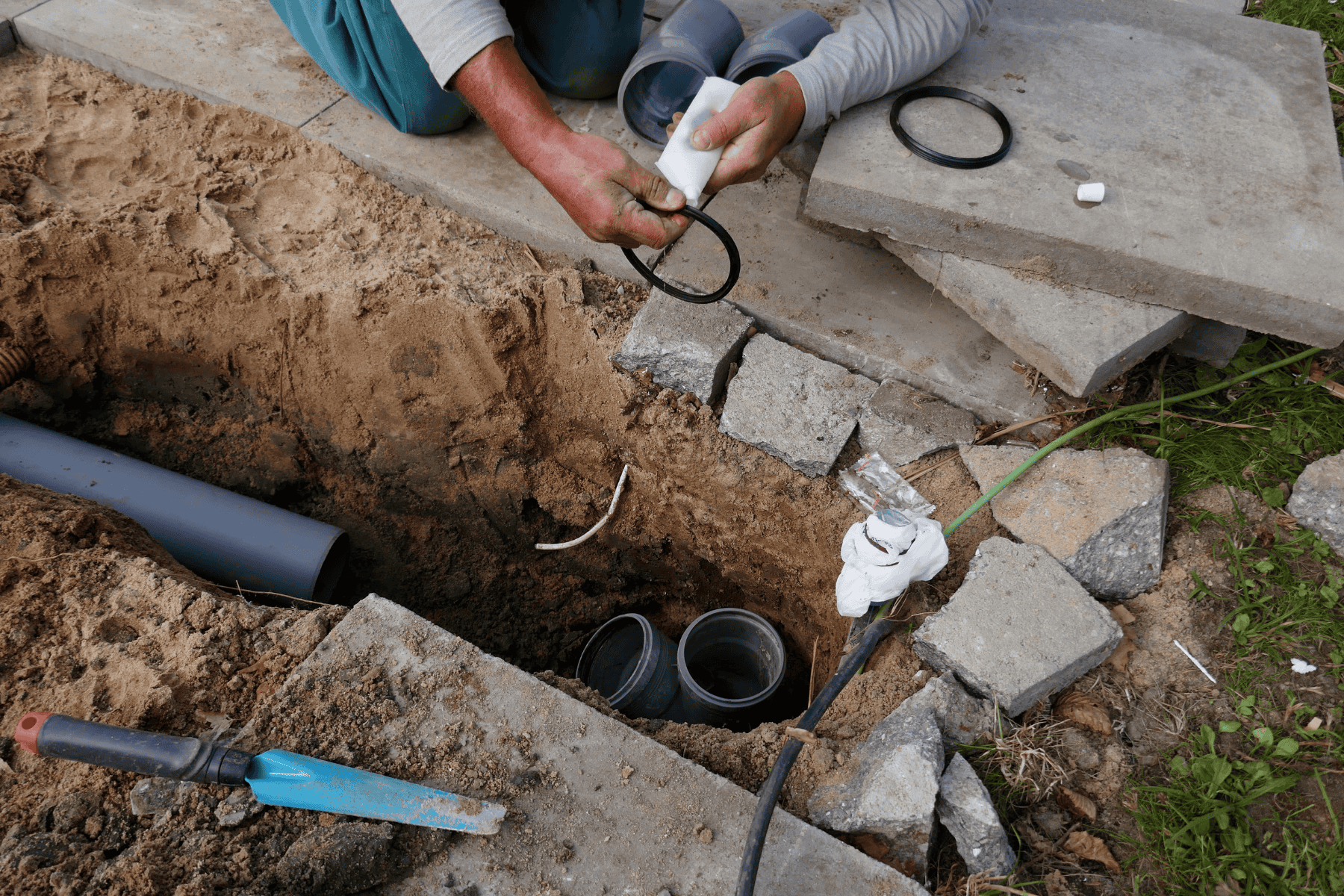 A person installs a black rubber seal onto a grey PVC pipe for a sewer repair, helping to keep the overall sewer repair cost manageable near an excavated trench.