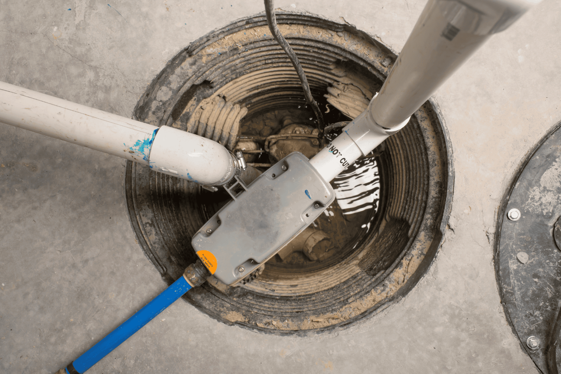 A circular utility access pit in a concrete floor containing white PVC piping, an electrical junction box, and a blue water line, often examined during sump pump troubleshooting.