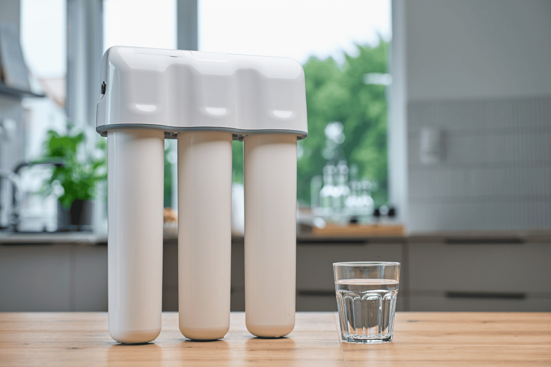 A three-stage under-sink water filtration system standing on a wooden table next to a glass of water, as discussed in our homeowner guide.